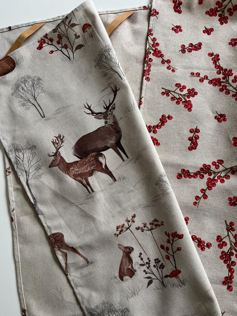 May include: Two beige fabric panels with nature-themed prints. One features deer, a rabbit, and trees in a winter scene. The other has a pattern of red berries on branches. Both have brown leather hanging loops.