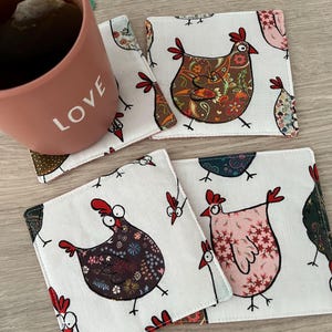 Könnte beinhalten: Vier quadratische Stoffuntersetzer mit weißem Hintergrund und einem sich wiederholenden Muster von skurrilen Hühnern in verschiedenen Farben und Mustern. Eine rosa Tasse mit dem Wort "LOVE" ist im Bild zu sehen. Für Heißgetränke.