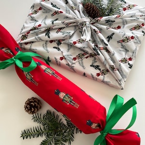 May include: A red fabric cracker and a white fabric-wrapped gift, both decorated with nutcracker soldier and evergreen branch patterns. Green ribbon accents the cracker. A pine cone and sprigs of greenery add to the festive holiday theme.