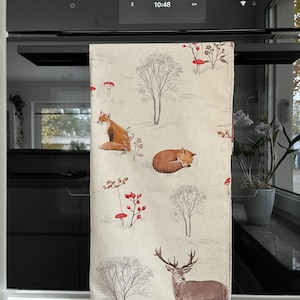 May include: A beige kitchen towel with a woodland pattern featuring foxes, deer, trees, and red mushrooms. The towel is hanging on a black oven.