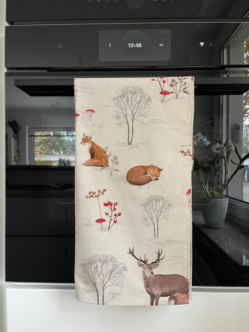May include: A beige kitchen towel with a woodland pattern featuring foxes, deer, trees, and red mushrooms. The towel is hanging on a black oven.