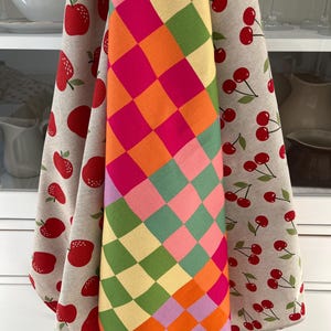 Könnte beinhalten: Drei Küchentücher mit farbenfrohen Designs. Ein Tuch hat ein Harlekinmuster in Pink, Orange, Grün und Gelb. Die anderen beiden Tücher haben Fruchtmuster: rote Äpfel und Kirschen auf hellbeigem Hintergrund.