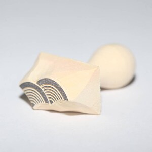 May include: A wooden stamp with a carved design of a wave pattern. The stamp is light brown and has a rounded handle.