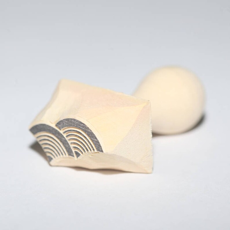 May include: A wooden stamp with a carved design of a wave pattern. The stamp is light brown and has a rounded handle.