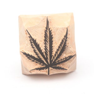 May include: A wooden stamp with a carved black cannabis leaf design. The stamp is a light brown color and has a square shape. The leaf is a detailed illustration of a cannabis plant.