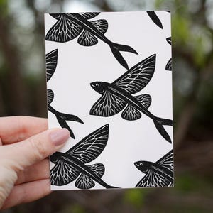Puede incluir: Una tarjeta blanca con un patrón repetido de peces voladores negros. Los peces tienen diseños detallados de alas y colas. La tarjeta está sostenida por una mano, con un fondo verde borroso.