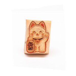 Könnte beinhalten: Ein Holzstempel mit einer lächelnden Maneki-neko-Katze, einem japanischen Glückssymbol, mit erhobener Pfote. Die Katze hat rote Akzente und hält eine Flasche mit japanischen Schriftzeichen. Der Stempel ist rechteckig.