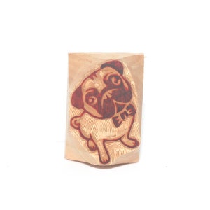 May include: A wooden stamp with a red and brown illustration of a pug wearing a bow tie.