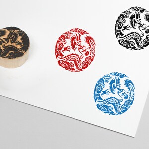 May include: A wooden stamp with a dragon design and three circular impressions in red, black, and blue. The dragon is surrounded by swirling clouds. The stamp is on a white surface.