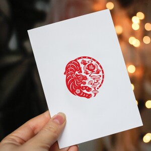 May include: White greeting card with a red tiger illustration in a circular design. The tiger is surrounded by stylized clouds and floral elements. The card is held in a person's hand.