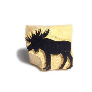 Puede incluir: Un sello con la silueta de un alce negro tallado en un bloque rectangular de madera de color claro. El alce tiene grandes astas y está de pie. El sello está diseñado para crear impresiones.