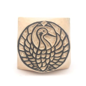 May include: A wooden stamp featuring a carved crane design within a circular frame. The crane is shown with outstretched wings, and the design is in black against the natural wood colour. The stamp is square-shaped.