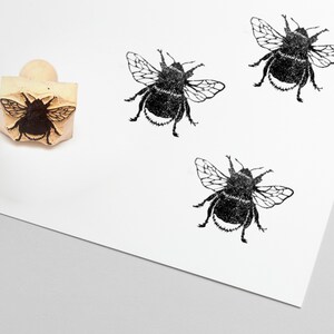 May include: A wooden stamp with a bumblebee design, next to three stamped impressions of the same bee. The bee stamps are black on a white background. The stamp has a rounded wooden handle.