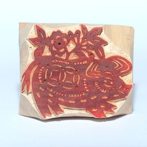 May include: A carved wooden stamp featuring a red pig design with floral accents. The stamp is rectangular and has a detailed, cut-out design. The pig is decorated with intricate patterns and flowers.