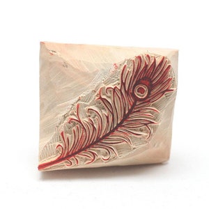 Puede incluir: Un sello de madera con un diseño de pluma de pavo real roja. El sello está tallado con detalles intrincados y está listo para ser utilizado para manualidades y proyectos de arte.