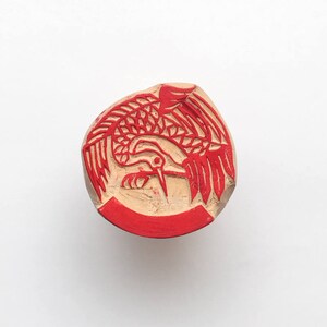 May include: Red wooden stamp with a detailed carving of a bird with its wings spread out.