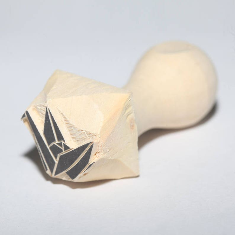 May include: A wooden stamp with a black origami crane design. The stamp is shaped like a rounded rectangle with a pointed end.