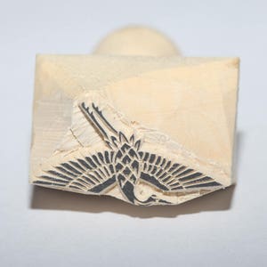 May include: A wooden stamp with a black ink design of a stylised crane with its wings spread.