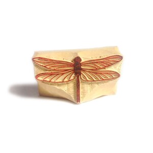 Peut inclure: Un tampon en bois avec un motif de libellule rouge. Le tampon est sculpté dans un bois brun clair et a une surface légèrement texturée.