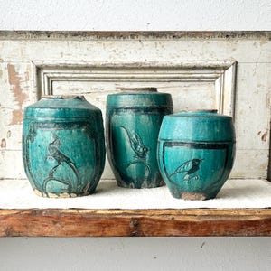 May include: Three antique turquoise ceramic jars with black bird illustrations. The jars vary in size and shape, with a cylindrical design and lids. The bird artwork is framed by a black border, adding a decorative touch to the aged pottery.