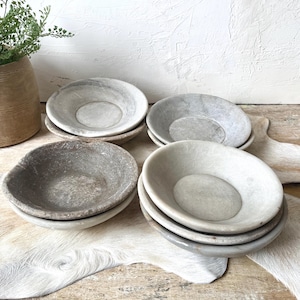 May include: A collection of four shallow stone bowls in varying shades of gray, beige, and off-white. Each bowl features a smooth, rounded interior and a textured, natural stone finish, stacked in two pairs on a light-colored surface.