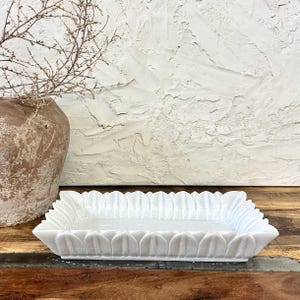 May include: A rectangular white marble tray with a scalloped edge design. The tray is placed on a wooden surface, with a textured white wall in the background. A brown vase with dried branches is on the left.
