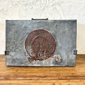 May include: A rectangular metal box with a tarnished, weathered appearance. A circular, embossed emblem with a figure and text is centered on the front. The box has a handle and hinges, suggesting it may be a vintage storage container.