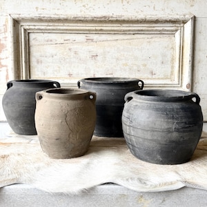 May include: A collection of four rounded earthenware pots featuring two small handles on each side. Three of the pots are dark charcoal gray with a matte finish, while one central pot is a lighter, weathered beige with a textured surface.