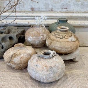 May include: A collection of small, antique ceramic vases in various shapes and sizes. The vases are primarily beige and brown, with some featuring decorative patterns. One vase is green. The vases are arranged on a textured surface.