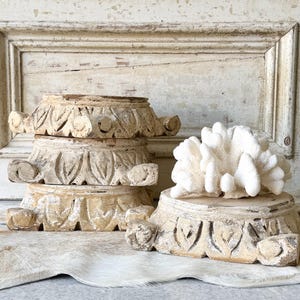May include: A stack of three circular, light-brown wooden pedestals featuring carved leaf-like patterns and a weathered finish. A fourth identical pedestal sits to the right, topped with a large, white, branching coral specimen.