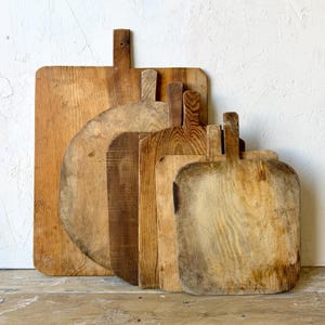 May include: A collection of five wooden cutting boards of varying shapes and sizes, each featuring a distinct grain pattern and a handle at the top. The boards are arranged in a layered formation, displaying light to dark brown wood tones.