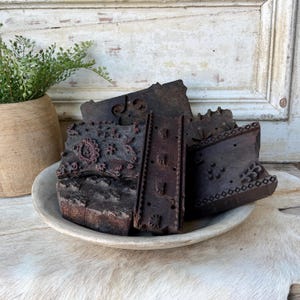 May include: A collection of dark brown wooden printing blocks with intricate carved patterns, including floral and geometric motifs, arranged inside a shallow, light-colored circular bowl. The blocks feature various shapes and textures.