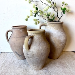 May include: Three weathered terracotta jugs with a matte, earthy finish and single side handles. The vessels feature varying sizes and organic, textured surfaces in shades of tan and beige. A delicate branch with small white flowers sits inside.