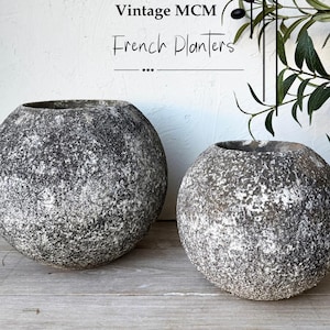 May include: Two spherical planters with a rough, textured surface in shades of dark gray and white. The text "Vintage MCM French Planters" is visible in the upper portion of the image, accompanied by a graphic of an olive branch.