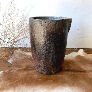 May include: A cylindrical vessel with a rough, textured surface featuring a dark brown and black mottled finish. The container has a thick rim and a slightly tapered base, showcasing a weathered appearance with visible variations in color tone.