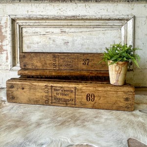 May include: Two weathered wooden crates with the text "HART & HERTEL" and numbers 37 and 69. A small potted plant with green foliage sits on top of the crates. The crates are made of aged wood.