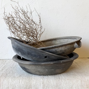 May include: Two stacked, shallow, dark gray ceramic bowls with a matte, weathered finish. The top bowl features a small side handle and a circular hole near the rim. A dry, brown, leafless branch is placed inside the upper bowl, extending upward.