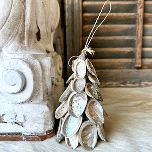 May include: A handcrafted Christmas tree ornament made from iridescent abalone shells. The tree-shaped decoration is strung together with natural twine, showcasing the shells' pearlescent colors. The ornament is displayed on a textured, light-colored surface.