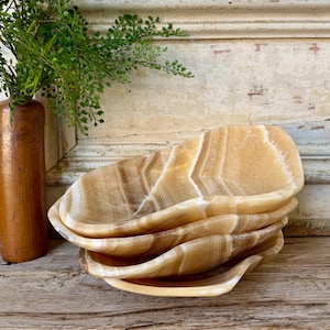 May include: A stack of four oval-shaped, amber-colored onyx bowls. The bowls have a natural, veined pattern with white and brown streaks. The bowls are stacked on a wooden surface, with a vase of greenery in the background.
