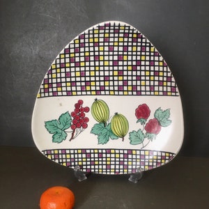 Op de afbeelding: Een vintage, driehoekige keramische plaat met een witte basis en een zwart, geel en paars geruit patroon. De plaat heeft een geschilderd ontwerp van rode bessen, kruisbessen en frambozen. Een kleine sinaasappel ligt voor de plaat.
