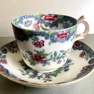 Könnte beinhalten: Ein Vintage-Tee-Set mit Tasse und Untertasse. Die weiße Porzellantasse und Untertasse haben ein Blumendesign mit rosa und roten Blumen, grünen Blättern und blauen Akzenten. Die Tasse hat einen roten Griff.