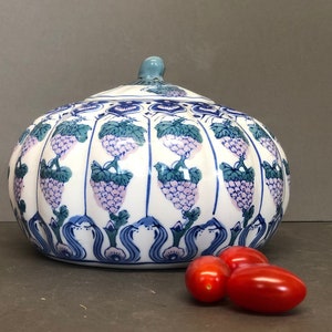 Puede incluir: Un tarro de cerámica blanca con tapa, decorado con un diseño floral azul y verde con uvas. El tarro tiene forma de calabaza y tiene una tapa redondeada. El tarro está sobre una superficie gris con tres tomates rojos delante.