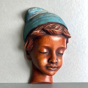 May include: A wooden sculpture of a person's head with closed eyes, wearing a blue and white hat. The sculpture is carved in a realistic style and has a warm brown colour.