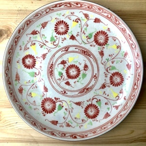 May include: A white ceramic plate with a red and yellow floral design. The plate has a raised rim and a black border.
