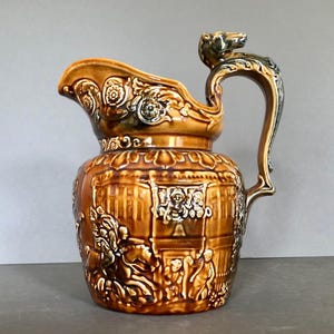 Könnte beinhalten: Braune Keramikkrug mit aufwendigen Reliefdetails. Der Krug hat einen dekorativen Griff mit einem geformten Tierkopf und einen Ausguss mit floralen Akzenten. Der Körper des Kruges zeigt Szenen mit Figuren und architektonischen Elementen.