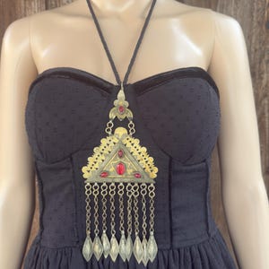 Puede incluir: Un vestido negro con un collar decorativo. El collar presenta un colgante triangular dorado con detalles rojos y colgantes plateados. El vestido tiene un corpiño sin tirantes y una falda fluida.