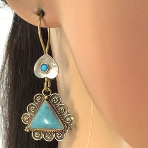 Puede incluir: Un solo pendiente con un gancho dorado y una parte superior plateada en forma de corazón con una pequeña piedra turquesa. La parte inferior presenta una piedra turquesa triangular rodeada por un marco decorativo dorado.