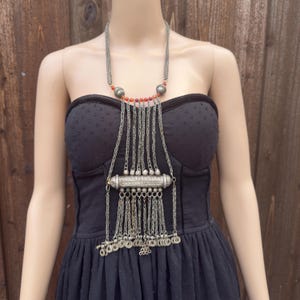 May include: Silver-toned statement necklace with a layered design. The necklace features a central cylindrical pendant, multiple chain dangles, and red bead accents. The necklace is displayed on a mannequin wearing a black dress.
