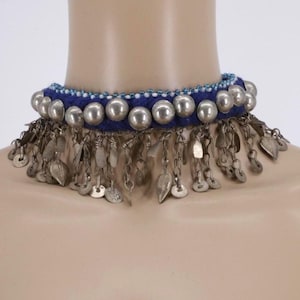 Puede incluir: Un collar de gargantilla de tela azul con adornos de metal plateado. El collar presenta grandes tachuelas redondas de metal y colgantes con forma de hoja y moneda. El borde superior está adornado con pequeñas cuentas blancas y azules.