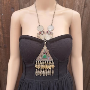 Puede incluir: Collar plateado con un colgante triangular y detalles de monedas. El colgante presenta una piedra verde y adornos coloridos. El collar se muestra sobre un vestido negro.